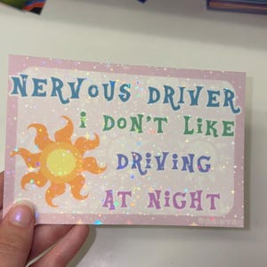 Puede incluir: Una pegatina rectangular con acabado hologr&aacute;fico de estrellas sobre un fondo rosa claro. Presenta un gr&aacute;fico de sol amarillo y naranja y el texto: NERVOUS DRIVER I DON'T LIKE DRIVING AT NIGHT en letras azules, verdes y moradas.