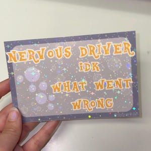 Puede incluir: Una pegatina rectangular con fondo morado oscuro que presenta un patr&oacute;n hologr&aacute;fico de estrellas y burbujas. El texto dice en letras naranjas con borde blanco: NERVOUS DRIVER IDK WHAT WENT WRONG. La pegatina tiene un acabado brillante.