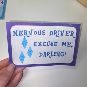 Pu&ograve; includere: Un adesivo rettangolare con bordo viola e centro bianco che riporta il testo blu "NERVOUS DRIVER EXCUSE ME, DARLING!". Il design include forme di diamanti blu e una finitura olografica che riflette la luce con piccoli motivi a stella.