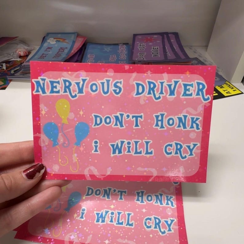Pu&ograve; includere: Un adesivo rettangolare rosa con finitura olografica che presenta il testo blu "NERVOUS DRIVER DON'T HONK I WILL CRY" e illustrazioni di tre palloncini in giallo e blu. Il design include piccoli dettagli a forma di stella e coriandoli.