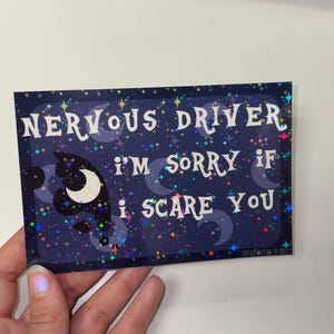 Puede incluir: Una pegatina rectangular con fondo azul oscuro que presenta un patr&oacute;n hologr&aacute;fico de estrellas y lunas crecientes. El texto dice: NERVOUS DRIVER I'M SORRY IF I SCARE YOU en letras blancas estilizadas. El dise&ntilde;o incluye peque&ntilde;as nubes oscuras.