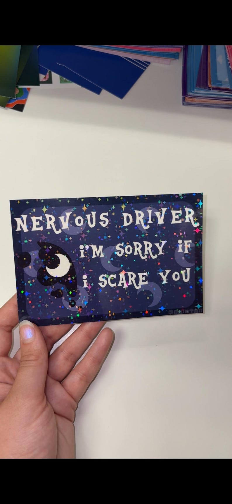 Puede incluir: Una pegatina rectangular con fondo azul oscuro que presenta un patr&oacute;n hologr&aacute;fico de estrellas y lunas crecientes. El texto dice: NERVOUS DRIVER I'M SORRY IF I SCARE YOU en letras blancas estilizadas. El dise&ntilde;o incluye peque&ntilde;as nubes oscuras.