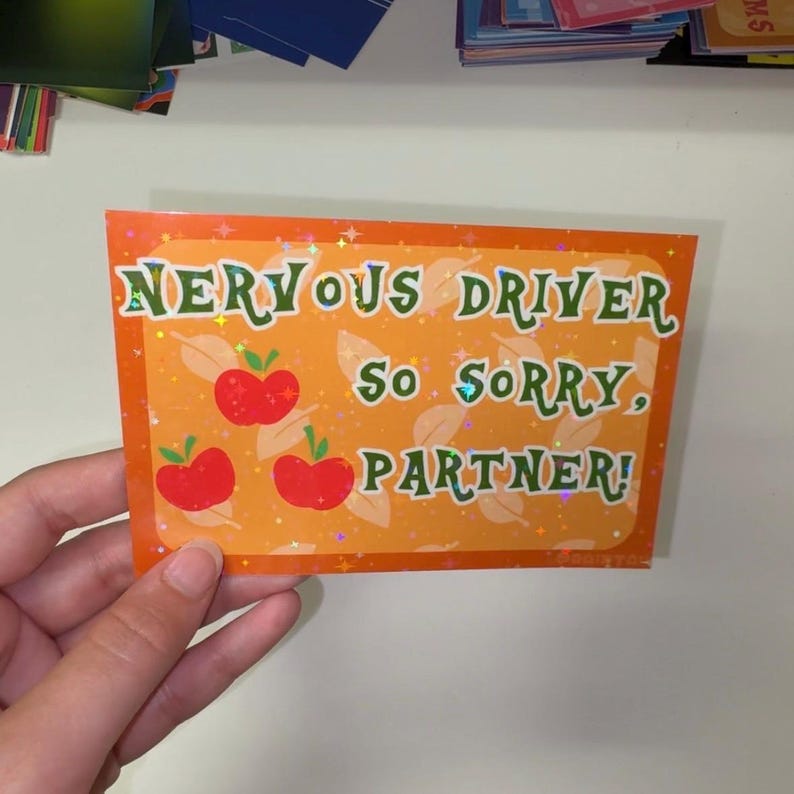 Pu&ograve; includere: Un adesivo rettangolare arancione con un motivo a stelle olografiche che presenta il testo verde NERVOUS DRIVER SO SORRY, PARTNER! e tre illustrazioni di mele rosse con foglie verdi. L'adesivo ha un bordo arancione scuro e uno sfondo arancione chiaro.