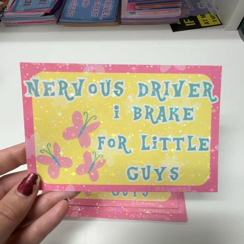 Pu&ograve; includere: Un adesivo rettangolare con centro giallo e bordo rosa caratterizzato da un motivo a stelle olografiche. Il testo NERVOUS DRIVER I BRAKE FOR LITTLE GUYS &egrave; scritto in blu, accompagnato da tre illustrazioni di farfalle rosa sulla sinistra.
