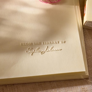 Peut inclure: Une étiquette de livre couleur crème avec le texte en relief FROM THE LIBRARY OF Sophie Johnson, ornée de trois œillets blancs et roses. Un stylo plume noir et une perforatrice noire sont placés à proximité de l'étiquette.