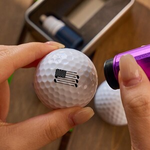 May include: A white golf ball with an American flag design is held near a purple golf ball marker. A small metal container and a blue bottle are in the background. The golf ball has a dimpled surface.