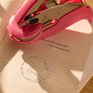 Peut inclure: Un tampon rose Trodat avec un mécanisme d'estampage en métal est posé sur un livre ouvert. Le livre contient du texte et un motif circulaire en relief avec l'inscription "FROM THE LIBRARY OF Tierney Arnold". Le tampon est marqué "UP-SIDE".