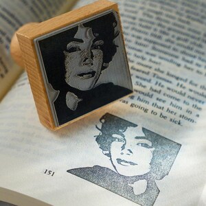 May include: A wooden stamp with a black and white image of a person's face. The stamp is on top of a book with the page number 151 visible.