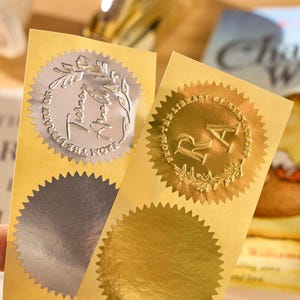 Peut inclure: Deux autocollants en feuille d'or et d'argent avec un bord dentelé. Les autocollants ont un design circulaire avec du texte et une couronne de laurier. Le texte sur les autocollants se lit "From the Library of..." et "RA". Les autocollants sont sur un fond jaune.