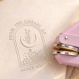 May include: A light pink metal embosser tool positioned next to a cream-colored paper surface featuring an embossed design. The design shows an arched frame containing a crescent moon, floral stems, and stars, with the text FROM THE LIBRARY OF ALICE GREEN.