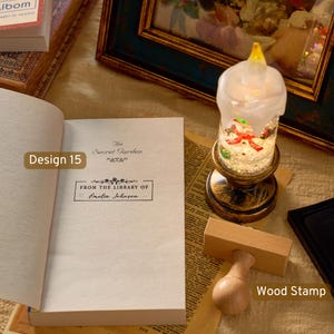 May include: A wooden stamp with a light-colored handle and a rectangular base, positioned next to an open book titled The Secret Garden. The book features a printed label that reads From the Library of Amelia Johnson, alongside a decorative candle.