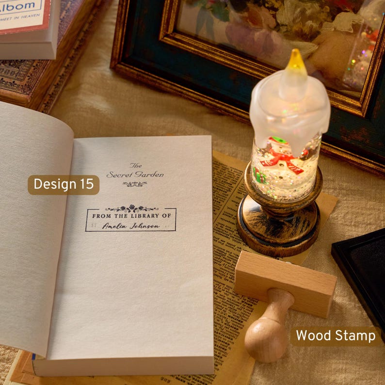 May include: A wooden stamp with a light-colored handle and a rectangular base, positioned next to an open book titled The Secret Garden. The book features a printed label that reads From the Library of Amelia Johnson, alongside a decorative candle.