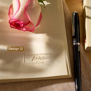 May include: A cream-colored book with an embossed rectangular frame containing the text "THIS BOOK BELONGS TO" and a name, accompanied by a pink and white rose and a black fountain pen. A small brown label displays the text "Design 12".