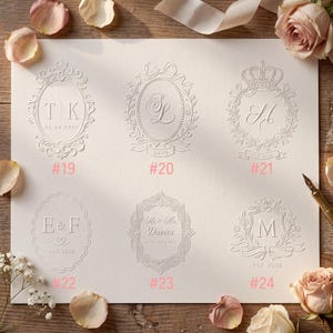 May include: A white card featuring six embossed monogram designs labelled #19 through #24. Each design includes unique decorative borders, initials, and dates. The card is surrounded by light pink rose petals and a fountain pen tip is visible.