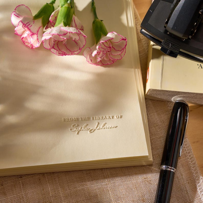 May include: A cream-colored bookplate featuring the embossed text FROM THE LIBRARY OF Sophie Johnson, decorated with three white and pink carnations. A black fountain pen and a black hole puncher are positioned near the bookplate.
