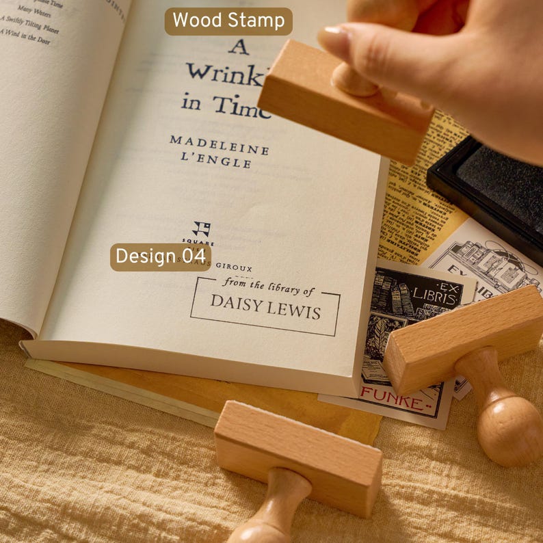 May include: A rectangular wooden stamp with a rounded handle, used to imprint the text from the library of DAISY LEWIS onto the open page of the book A Wrinkle in Time by Madeleine L'Engle, alongside other wooden stamps and ink pads.