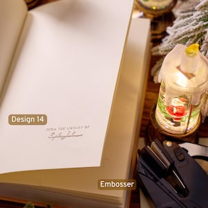 May include: An open book with a cream-colored page featuring a debossed impression that reads "FROM THE LIBRARY OF" followed by a cursive name. A black handheld embosser tool is positioned nearby on the surface next to a small illuminated lantern.