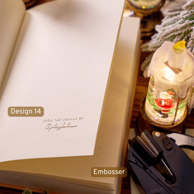 May include: An open book with a cream-colored page featuring a debossed impression that reads "FROM THE LIBRARY OF" followed by a cursive name. A black handheld embosser tool is positioned nearby on the surface next to a small illuminated lantern.