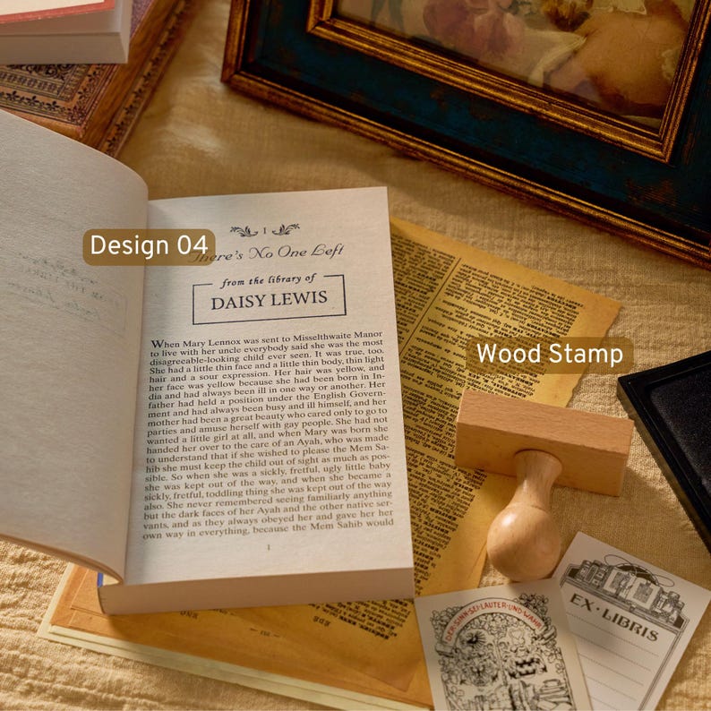 May include: A wooden rubber stamp with a rounded handle and a rectangular base, positioned next to an open book displaying the text "There's No One Left" and "from the library of DAISY LEWIS," alongside a small card featuring an "EX LIBRIS" illustration.