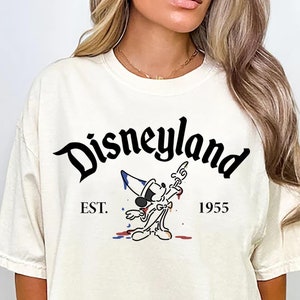 May include: A white t-shirt with a black graphic of Mickey Mouse in front of a paint splatter. The text "Disneyland" is above the graphic and "EST. 1955" is below the graphic.