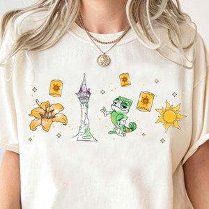 May include: A white t-shirt with a cartoon image of a chameleon, a tower, a flower, and a sun. The chameleon is green and has a surprised expression. The tower is tall and has a purple roof. The flower is yellow and has five petals. The sun is yellow and has a smiling face.