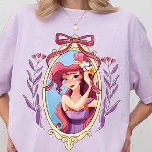 May include: A lavender t-shirt with a cartoon image of a woman with long red hair in a gold frame. The woman is wearing a purple dress and has a flower in her hair. The frame is decorated with a red bow and purple vines.
