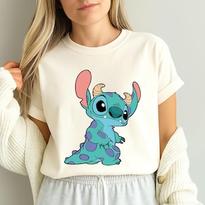 May include: A white t-shirt featuring a blue and purple cartoon character with large eyes and pink ears. The character is Stitch from the Disney movie Lilo & Stitch. The shirt is available in ivory.
