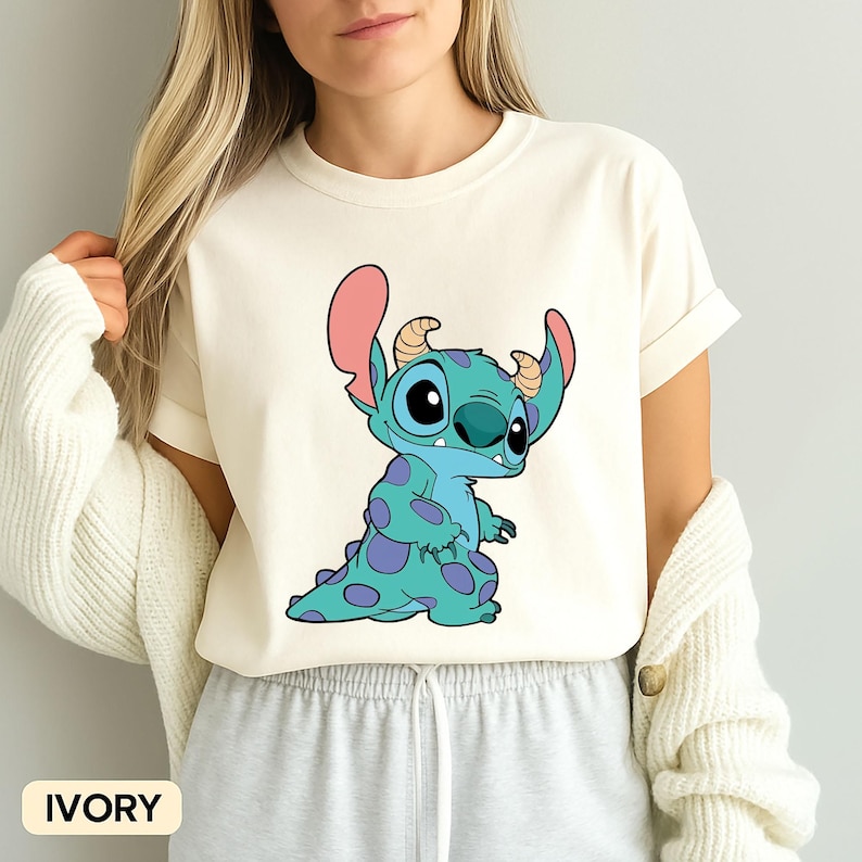 May include: A white t-shirt featuring a blue and purple cartoon character with large eyes and pink ears. The character is Stitch from the Disney movie Lilo & Stitch. The shirt is available in ivory.