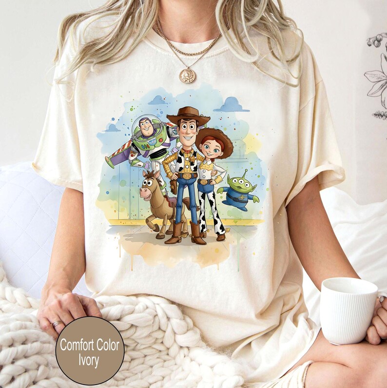 May include: Ivory-colored t-shirt featuring a watercolor illustration of characters from the animated film "Toy Story." The design includes Woody, Buzz Lightyear, Jessie, and other characters. The text "Comfort Color Ivory" is at the bottom.