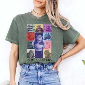 May include: A green t-shirt featuring a collage of Shrek faces in various colours. The text "IN MY SHREK ERA" is printed beneath the collage.
