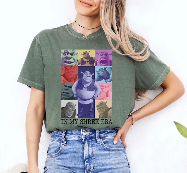 May include: A green t-shirt featuring a collage of Shrek faces in various colours. The text "IN MY SHREK ERA" is printed beneath the collage.