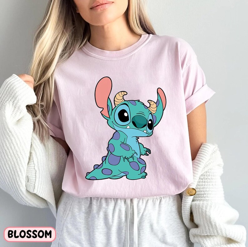 May include: A pink t-shirt featuring a cartoon blue Stitch character from the Disney film Lilo & Stitch. Stitch is wearing a blue and purple outfit with white eyes and a pink nose.