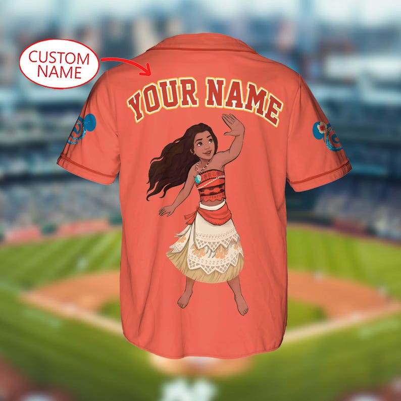 Pu&ograve; includere: Maglia da baseball color corallo con un personaggio dei cartoni animati. La parte posteriore della maglia mostra la scritta "YOUR NAME" in oro sopra il personaggio. Le maniche hanno disegni blu e bianchi. La maglia &egrave; su uno sfondo sfocato di campo da baseball.