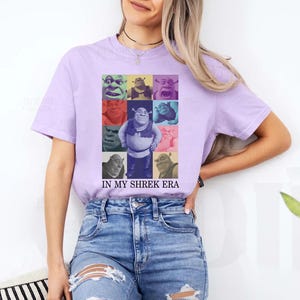 May include: A purple t-shirt featuring a collage of Shrek characters in various colours. The text "IN MY SHREK ERA" is printed beneath the collage.