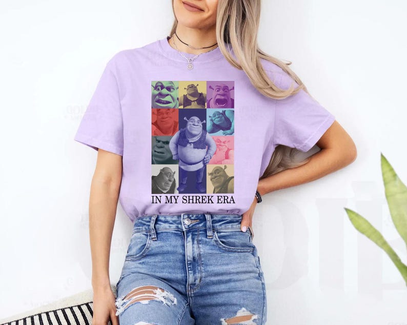 May include: A purple t-shirt featuring a collage of Shrek characters in various colours. The text "IN MY SHREK ERA" is printed beneath the collage.