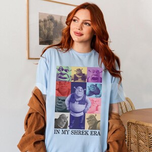 May include: Light blue t-shirt featuring a colourful collage of Shrek characters. The text "In My Shrek Era" is printed on the bottom of the shirt.