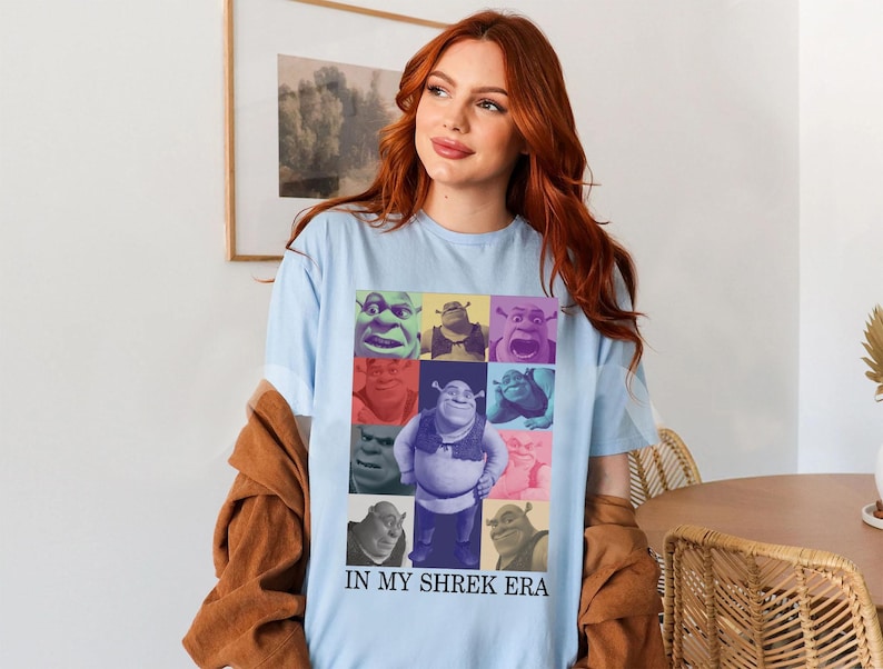 May include: Light blue t-shirt featuring a colourful collage of Shrek characters. The text "In My Shrek Era" is printed on the bottom of the shirt.