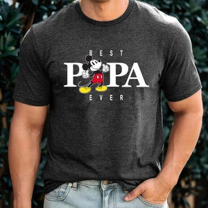 May include: A dark grey t-shirt with a white graphic of Mickey Mouse saying "Best Papa Ever".