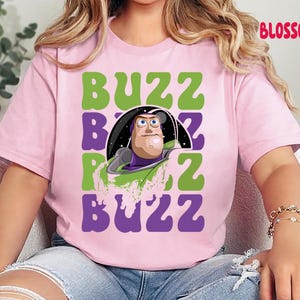 May include: A light pink t-shirt featuring a graphic of Buzz Lightyear from Toy Story. The design includes the word "BUZZ" in green and purple block letters, with Buzz's image in the center. The word "BLOSSOM" is printed in red on the top right.