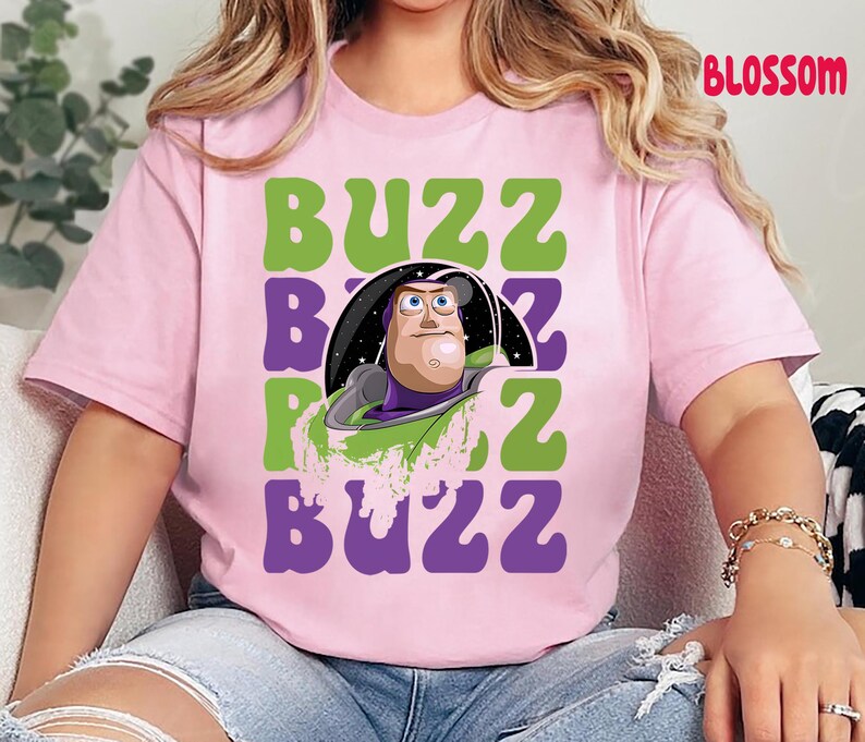May include: A light pink t-shirt featuring a graphic of Buzz Lightyear from Toy Story. The design includes the word "BUZZ" in green and purple block letters, with Buzz's image in the center. The word "BLOSSOM" is printed in red on the top right.