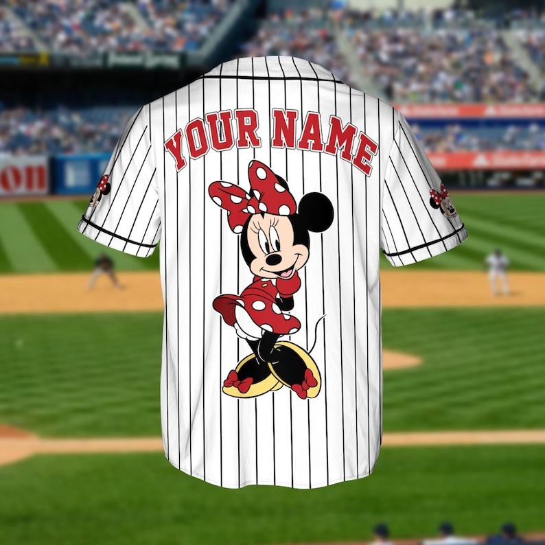 May include: White baseball jersey with black vertical stripes, featuring a Minnie Mouse graphic on the back. The jersey has the text "YOUR NAME" in red arched across the upper back. Minnie Mouse is wearing a red polka dot dress and bow.