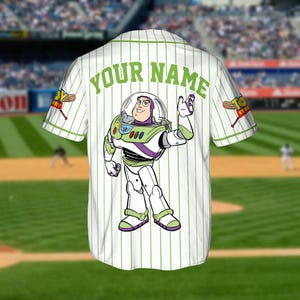 May include: White baseball jersey with green pinstripes and the text "YOUR NAME" across the back. Features a Buzz Lightyear graphic and "Toy Story" logo on the sleeve. The jersey is on display in a baseball stadium.