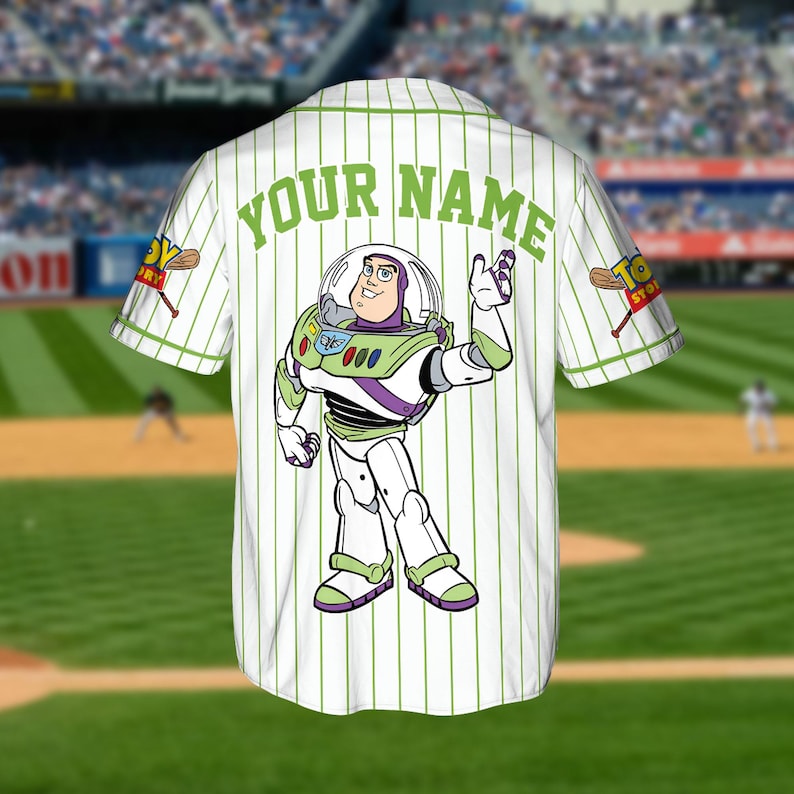May include: White baseball jersey with green pinstripes and the text "YOUR NAME" across the back. Features a Buzz Lightyear graphic and "Toy Story" logo on the sleeve. The jersey is on display in a baseball stadium.