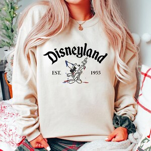 May include: A cream-colored sweatshirt with a black graphic of Mickey Mouse holding a paintbrush and a paint can. The text "Disneyland" is in black above the graphic and "EST. 1955" is in black below the graphic.