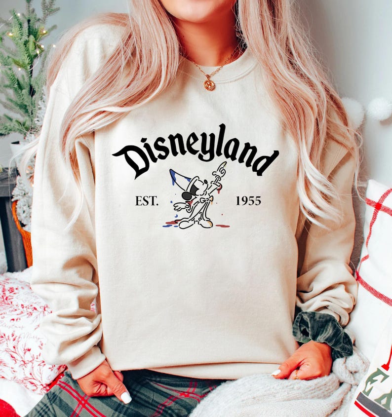 May include: A cream-colored sweatshirt with a black graphic of Mickey Mouse holding a paintbrush and a paint can. The text "Disneyland" is in black above the graphic and "EST. 1955" is in black below the graphic.