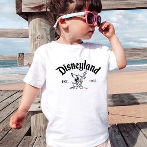May include: White t-shirt with a black and white illustration of Mickey Mouse wearing a sorcerer hat and holding a wand. The text "Disneyland" is in a black cursive font above the illustration. The text "EST. 1955" is below the illustration.
