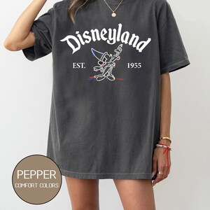 May include: A dark gray t-shirt with a white graphic of Mickey Mouse as a sorcerer, with the text "Disneyland" above and "EST. 1955" below.