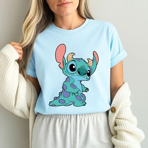 May include: A light blue t-shirt featuring a cartoon image of Stitch, a blue alien character with purple spots, from the Disney film Lilo & Stitch.