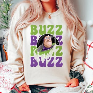 May include: Cream-colored sweatshirt featuring a graphic of Buzz Lightyear with the word "BUZZ" in green and purple. The design includes a cartoon image of Buzz Lightyear in his space suit. The sweatshirt is a casual, comfortable style.