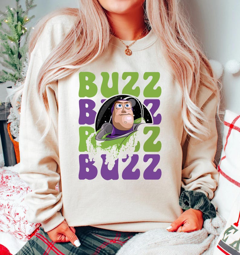 May include: Cream-colored sweatshirt featuring a graphic of Buzz Lightyear with the word "BUZZ" in green and purple. The design includes a cartoon image of Buzz Lightyear in his space suit. The sweatshirt is a casual, comfortable style.